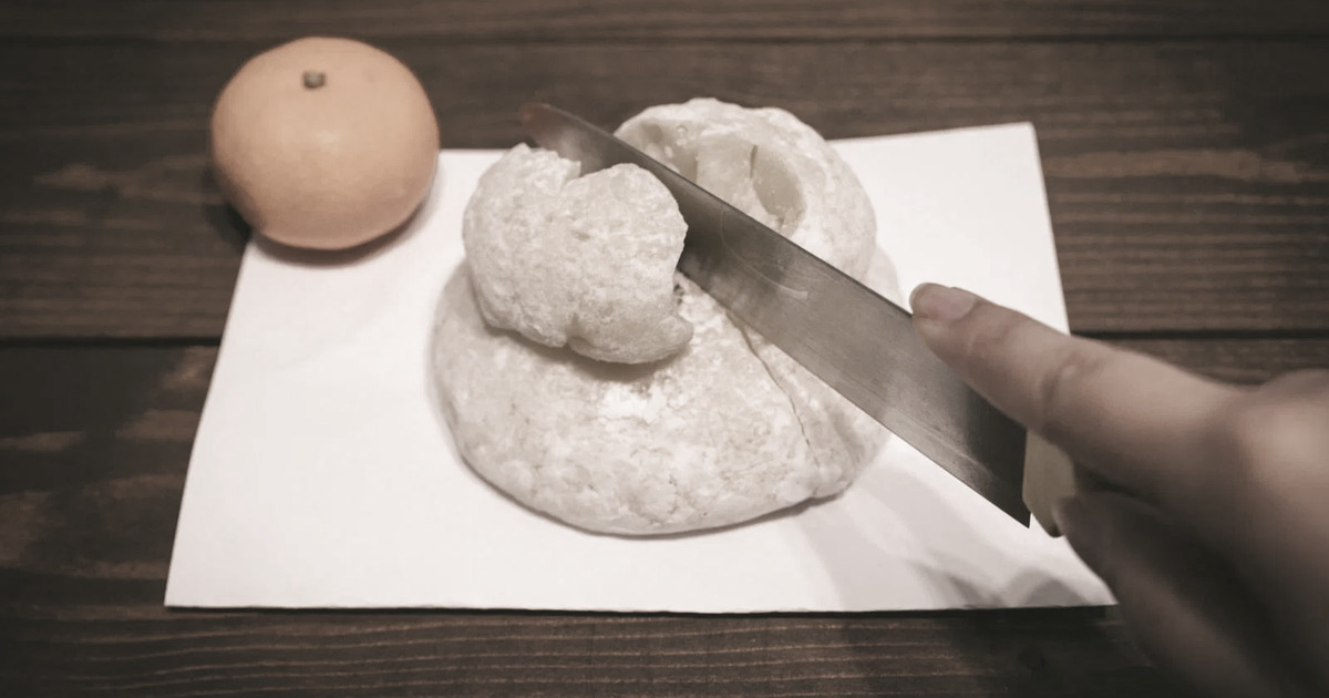 A hand holding a kitchen knife against a dried, cracked kagami mochi.